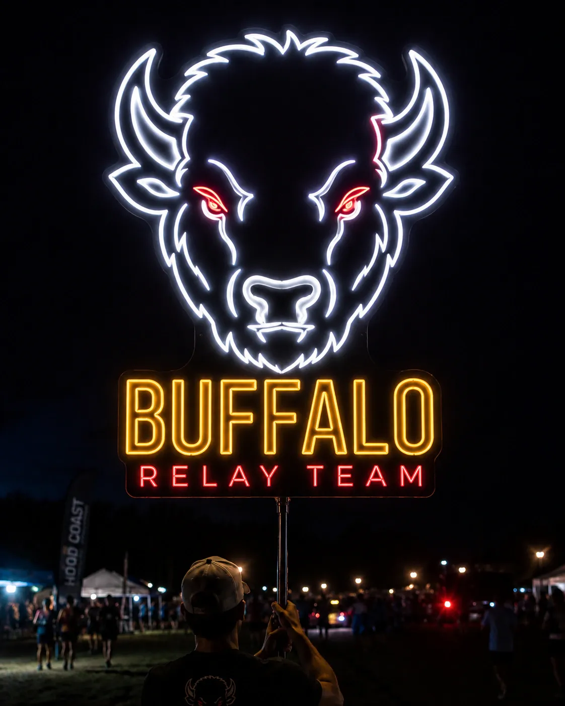 A lit neon buffalo head over the words Buffalo Relay Team, held up on a pole at night above a race crowd.