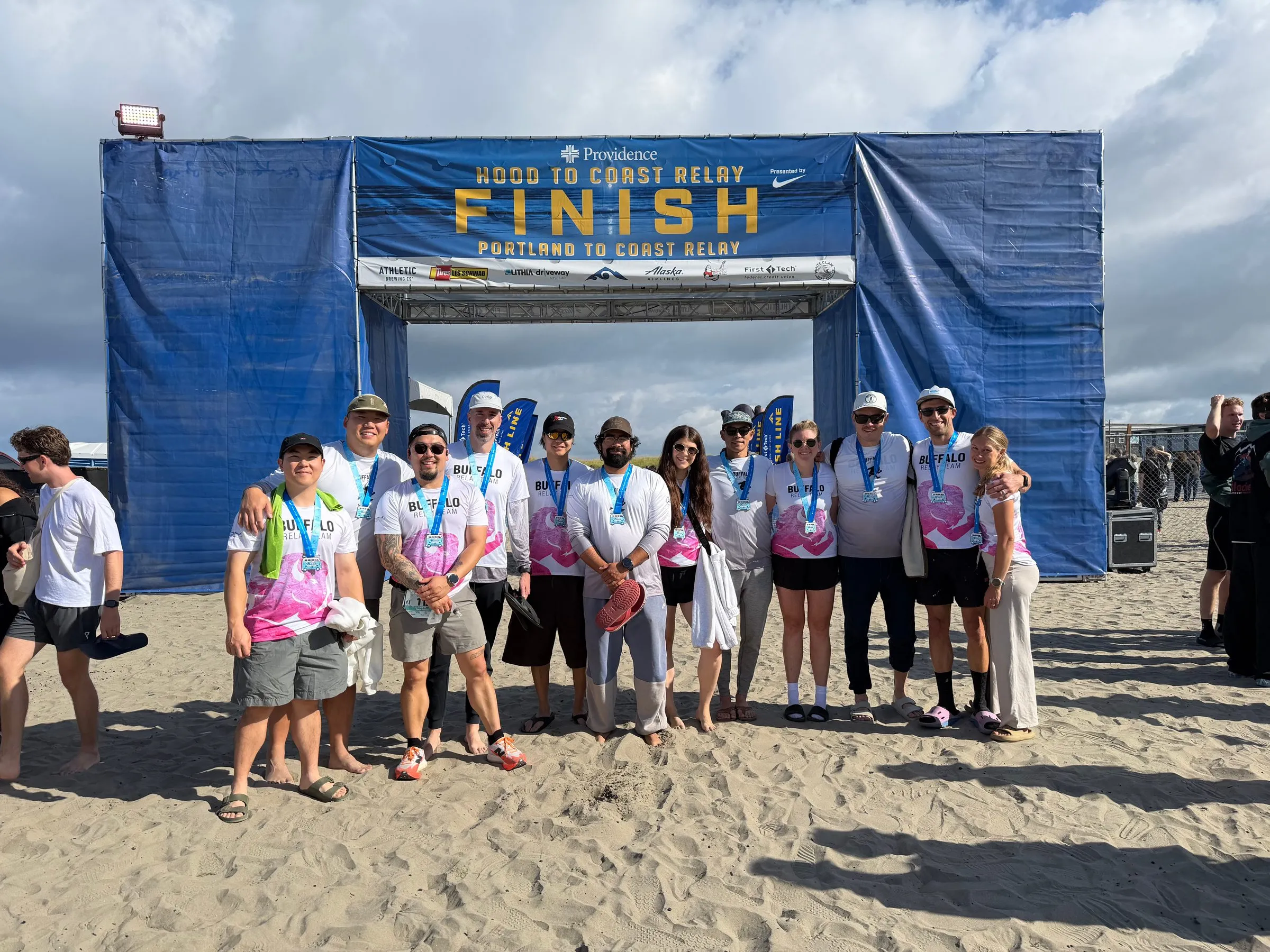 Twelve teammates in jerseys and finisher medals stand in a row on the sand beneath the Relay Finish arch.