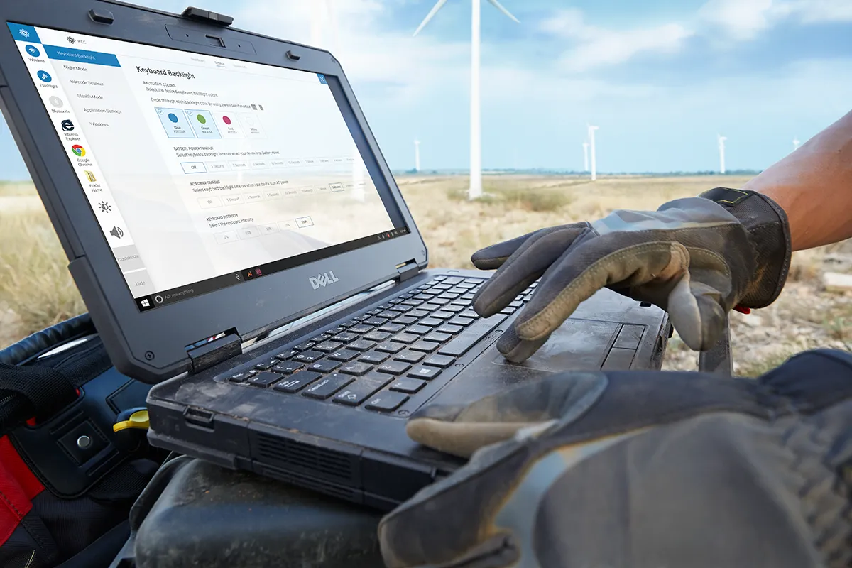 A gloved hand on a rugged Dell laptop outdoors near wind turbines, with Rugged Control Center on screen.