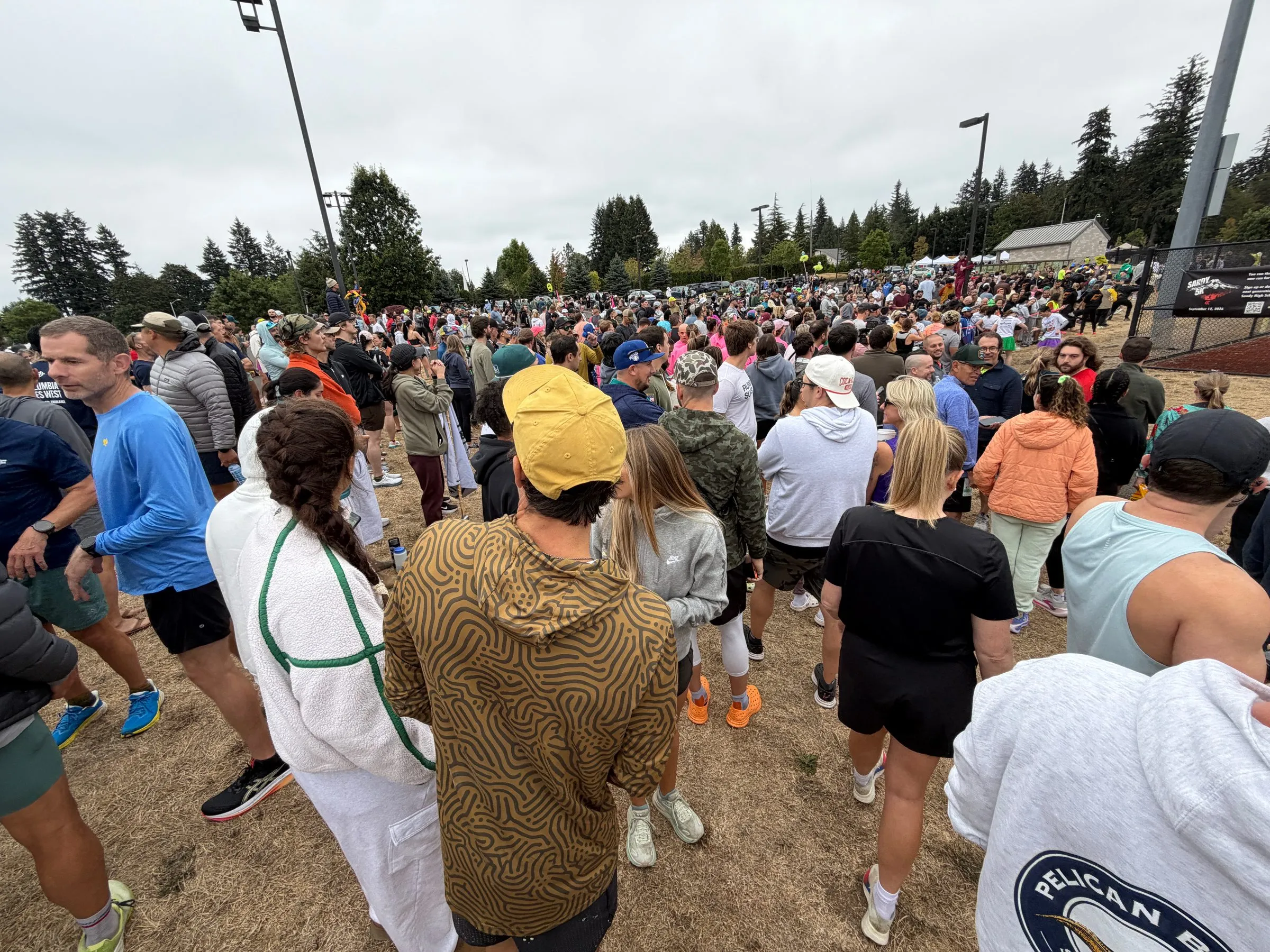 A dense crowd of runners and spectators on dry grass under an overcast sky, with evergreens and tents behind.