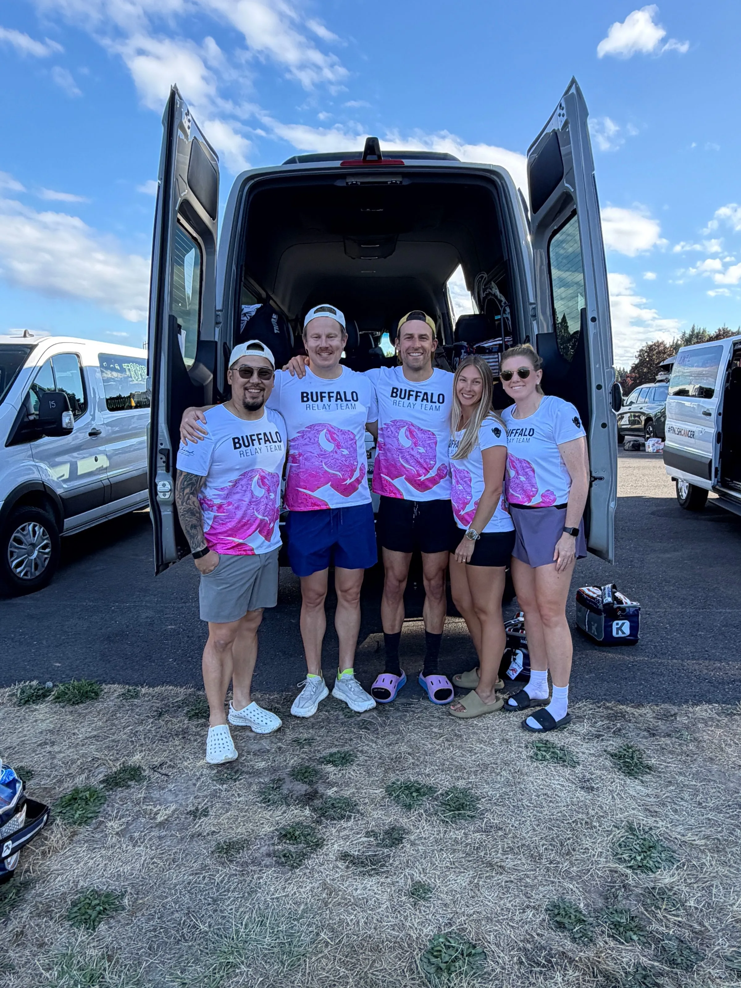 Five teammates in matching pink-and-white jerseys stand at the open rear doors of a van under a blue sky.