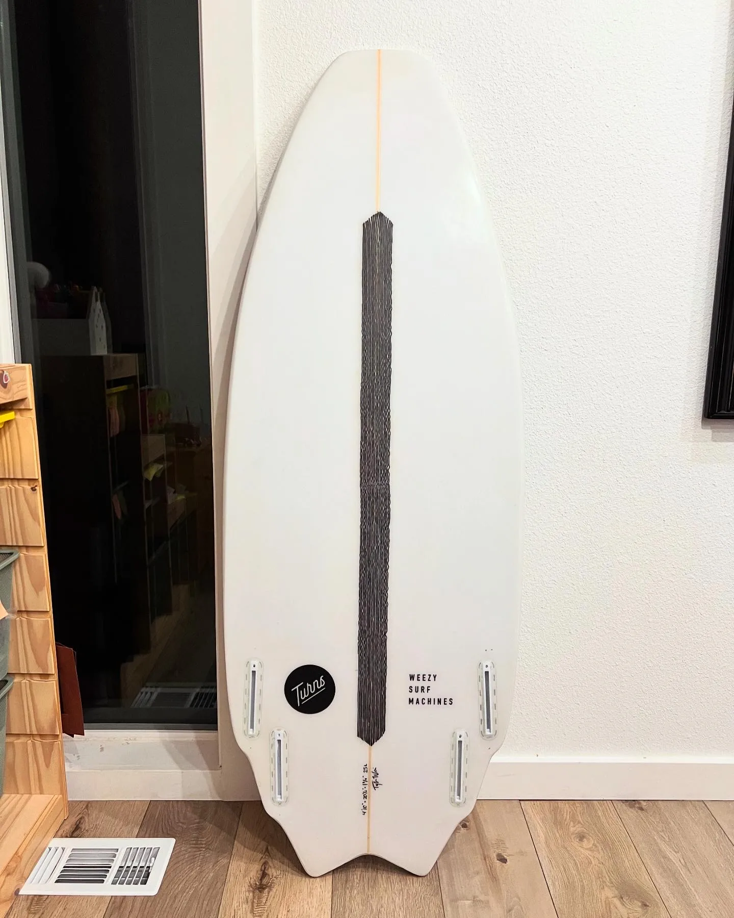 A white surfboard with a black carbon strip down the middle and five fin boxes, leaning against a wall.